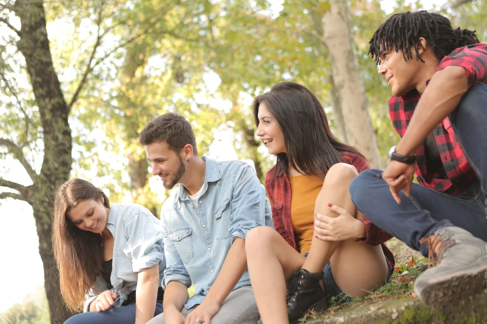 Four friends sit together on a sunny grassy slope in a park, smiling and chatting