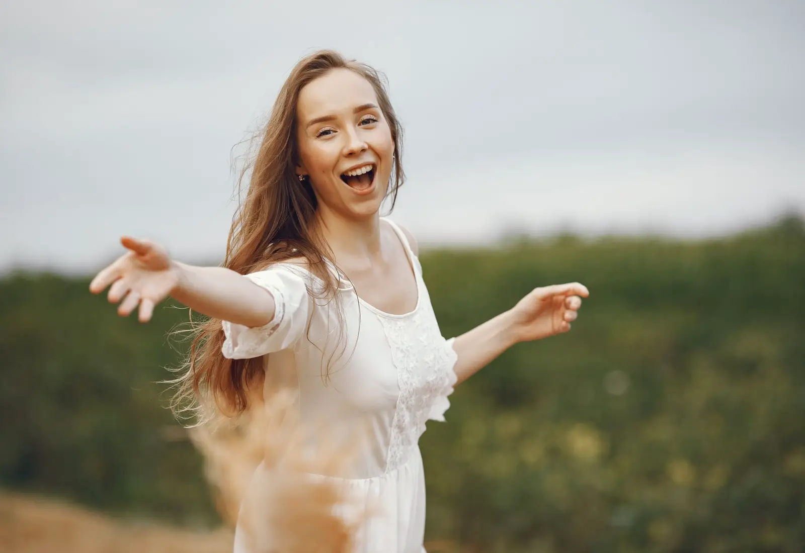A smiling woman in a white dress walks toward the camera with her arms open in a sunny green park