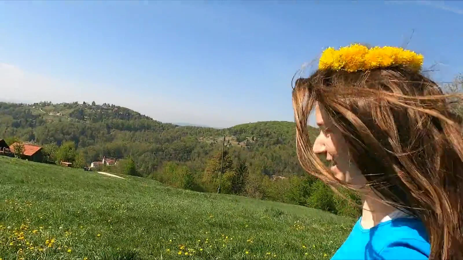 A smiling woman wearing a flower crown stands in a green hillside meadow under a bright blue sky