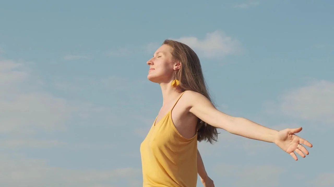 A smiling woman in a yellow top stands in the sunshine with her arms spread wide and her face lifted to a clear blue sky