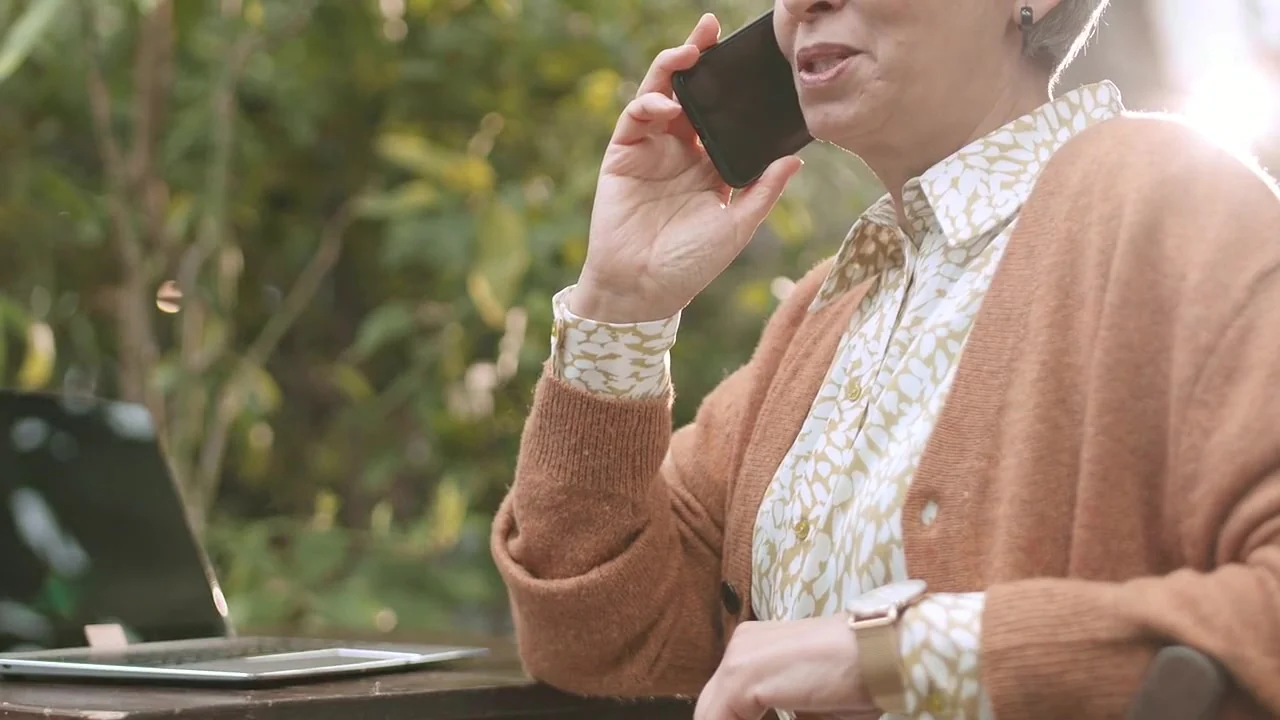 A smiling woman in a blouse and cardigan talks on the phone at an outdoor table in warm sunlight, her laptop open in front of her