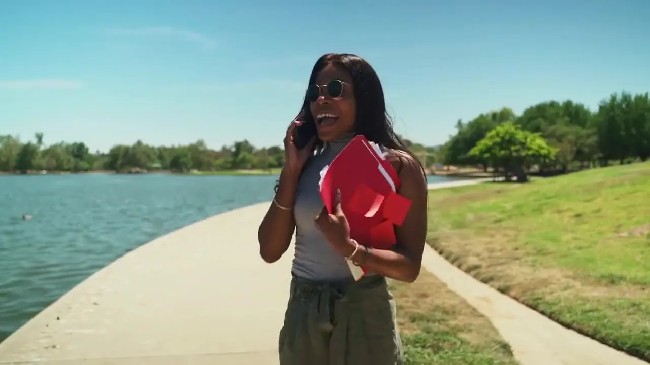 A smiling woman walks beside a sunny lake while talking on a phone call